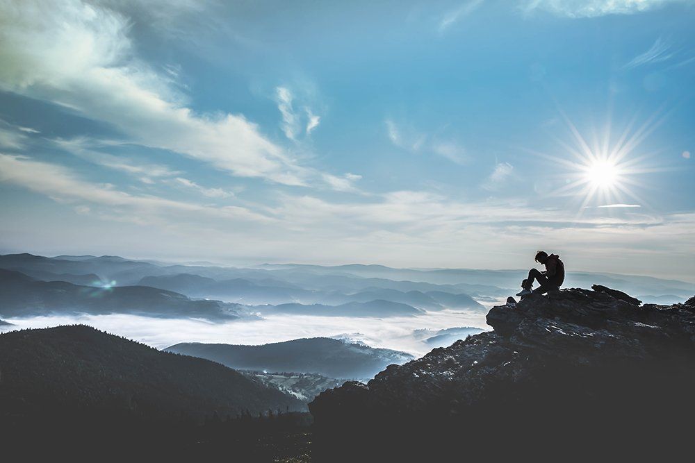 Silhouette of person sitting on mountain peak, overlooking misty valleys under a bright sun.