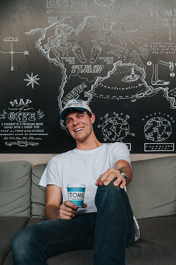 Man sitting, smiling, holding a coffee cup. Wearing white shirt and cap, against a world map mural.